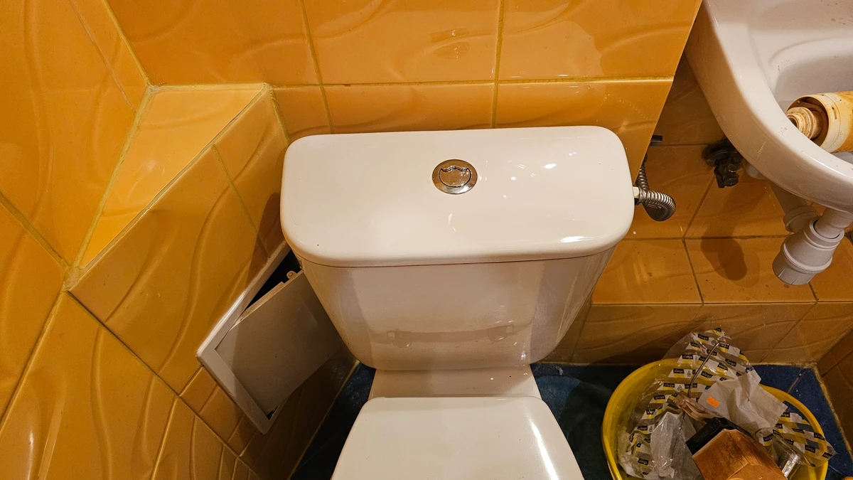 Biały kompakt WC z przyciskiem spłukującym na tle pomarańczowych płytek w bloku, obok umywalka i miska z nową armaturą spłuczki