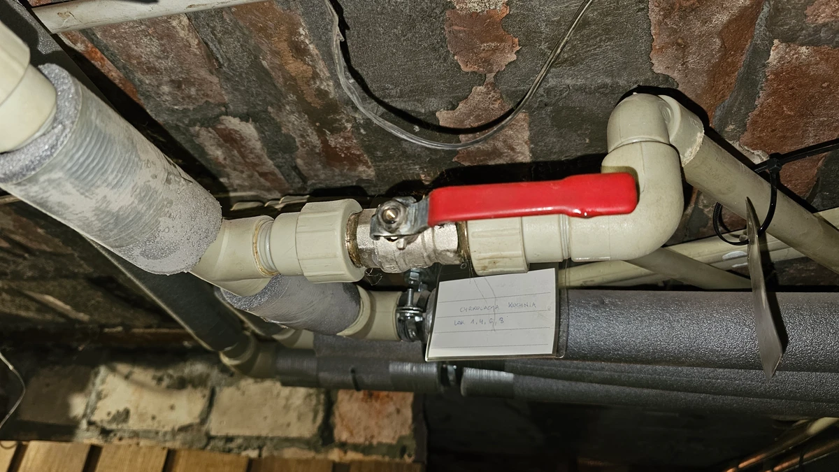 Mosiężny zawór kulowy z czerwoną dźwignią wpięty przez śrubunki w instalację z polipropylenu pod stropem piwnicy bloku