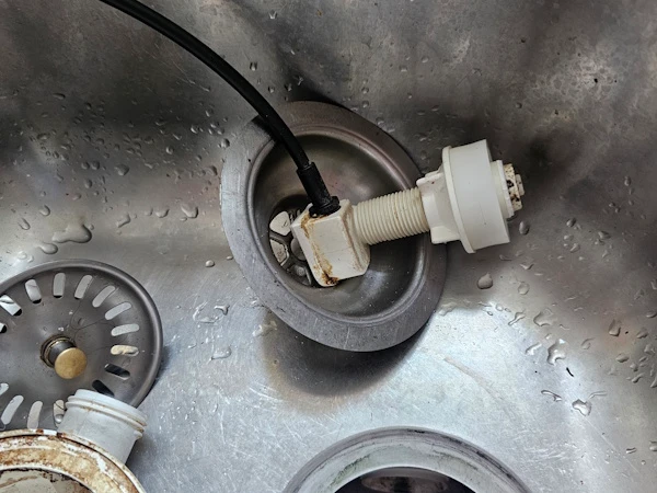 Nowa śruba mocująca sitko wkręcona w otwór odpływu zlewu, obok czarne cięgno mechanizmu